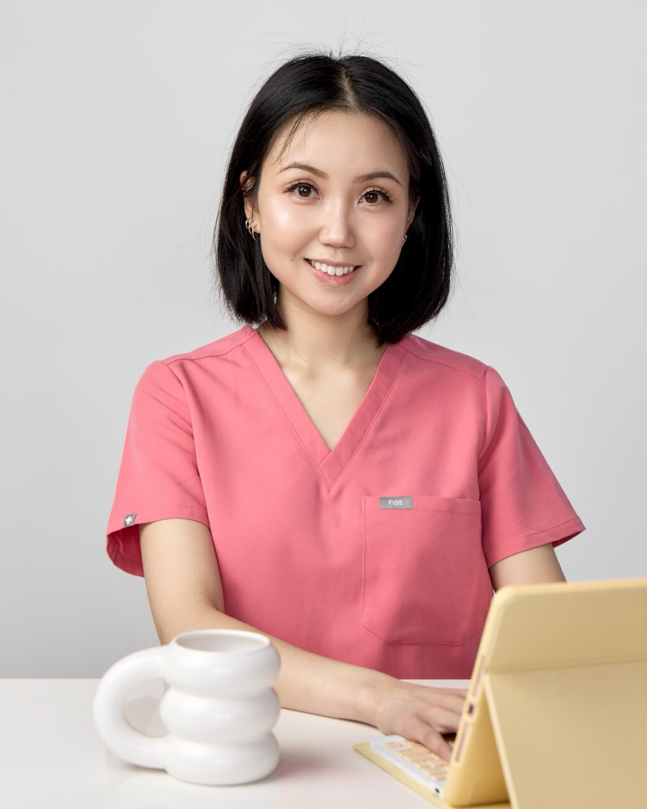 Dr. Chloe Yee, Vancouver naturopathic doctor, smiling in pink medical scrubs with short black hair, seated at a table with a white coffee mug and yellow iPad.