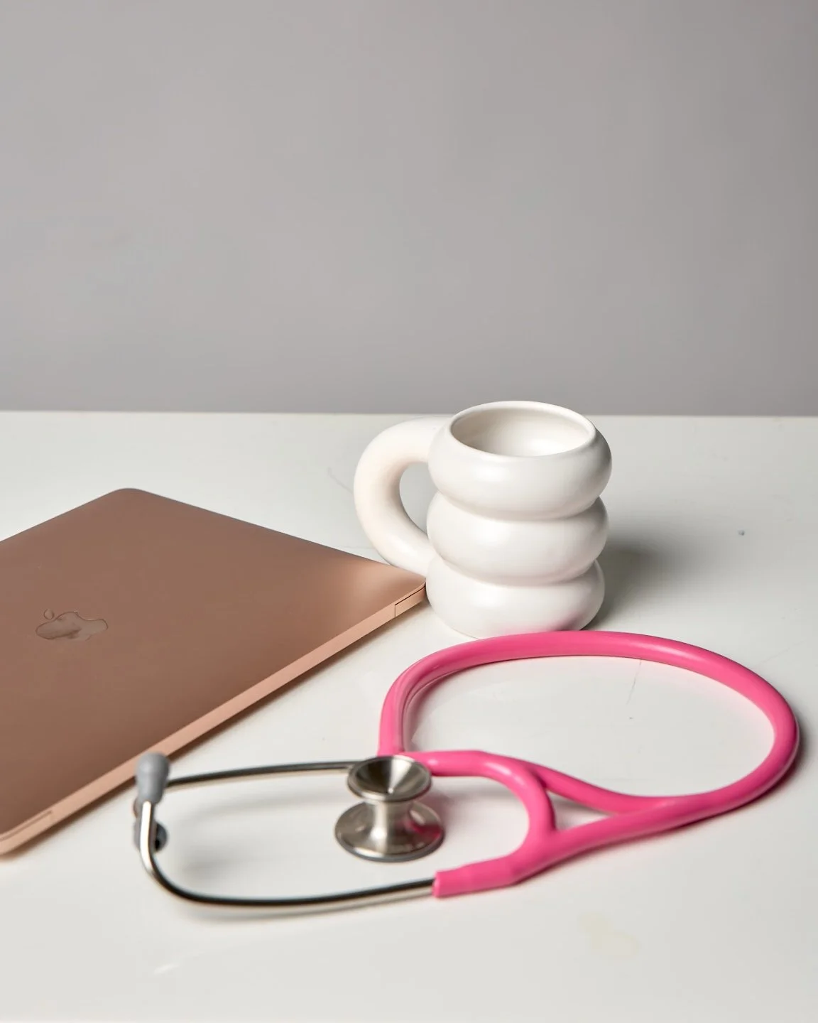 A pink stethoscope, a closed rose gold MacBook, a white ceramic mug with a stacked design, and a gray background on a white desk.