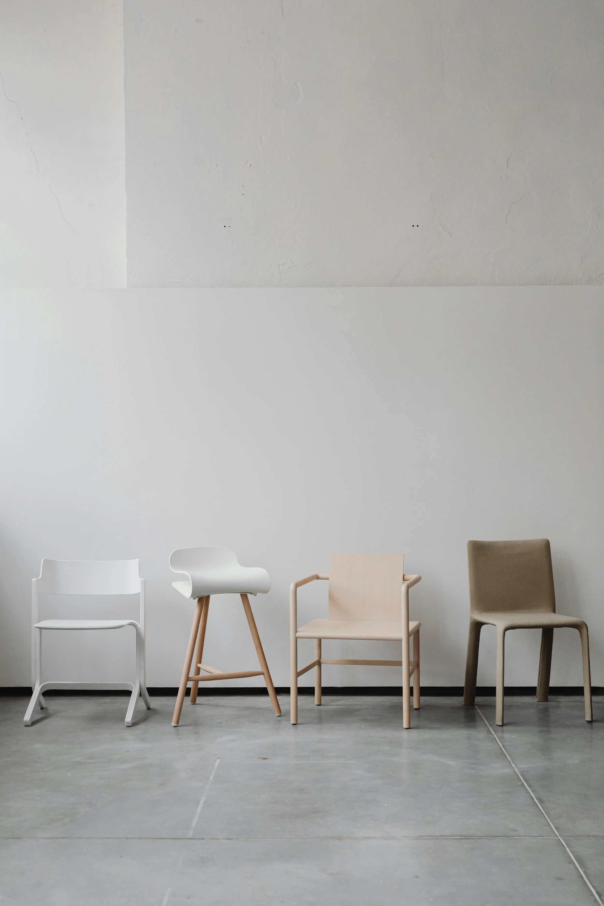 Four modern chairs lined up against a white wall in a minimalist setting, with a concrete floor.