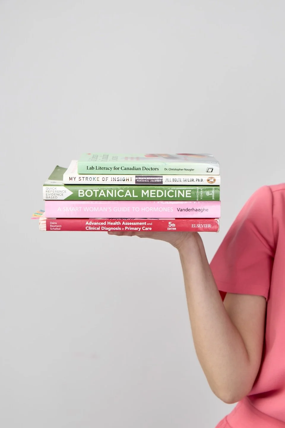 A person in pink scrubs holding a stack of five medical and health-related books against a plain gray background.