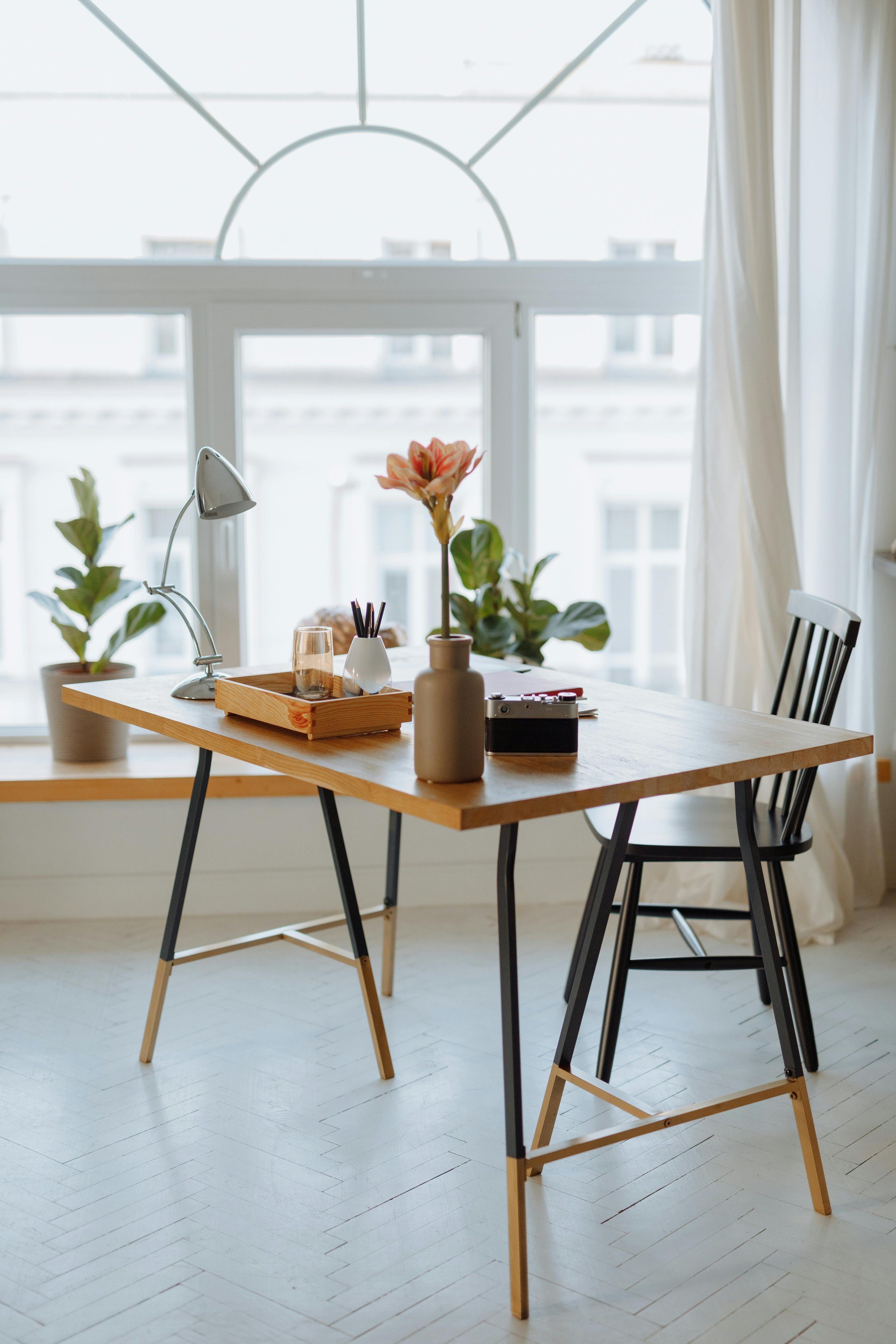 A bright workspace with a wooden desk, black chairs, and plants by a large window with white curtains, decorated with a bottle holding a pink flower, a desk lamp, and various small office supplies.