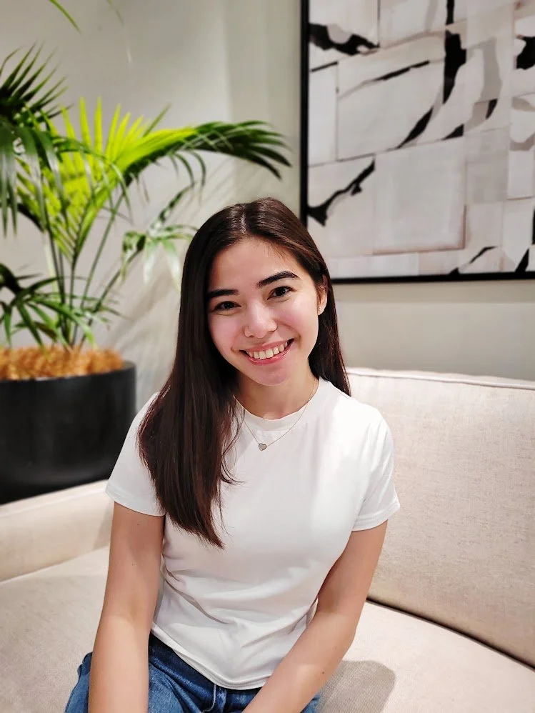 Young woman with long dark hair smiling, wearing a white t-shirt, sitting on a beige couch, with green plants and abstract artwork in the background.