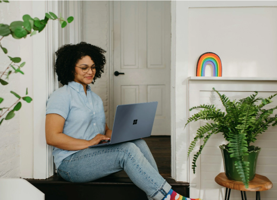 Woman sitting on the floor leaning against a wall, working on a silver laptop, with a large green potted fern nearby and a rainbow decoration on the wall.