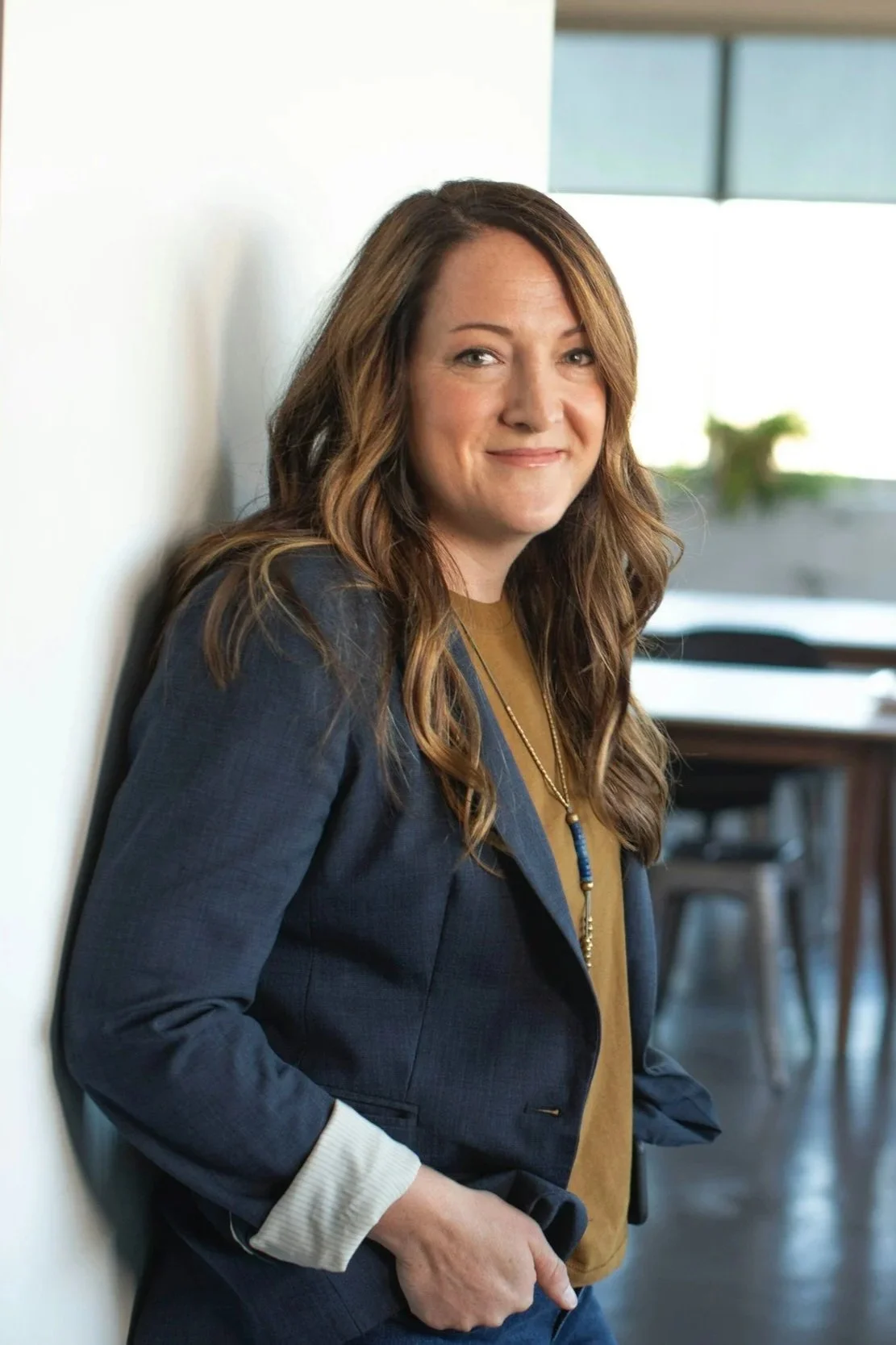 A woman with long wavy brown hair, wearing a navy blazer over a mustard shirt, smiling and leaning against a white wall in an office setting with large windows and chairs in the background.