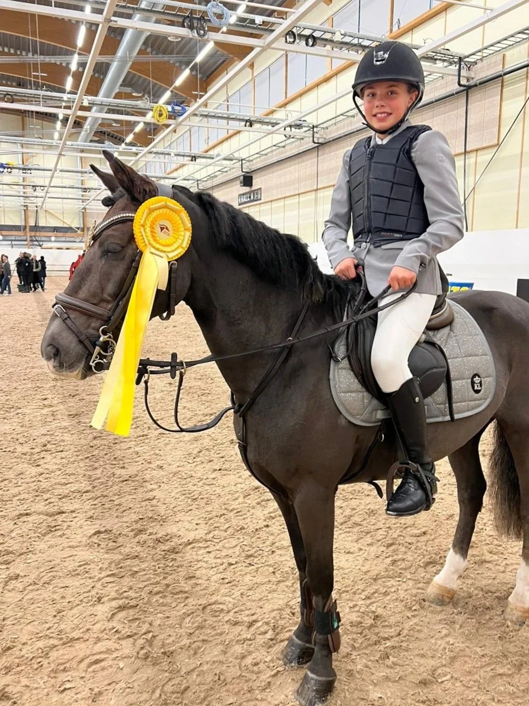 A young rider dressed in equestrian attire sitting on a dark brown horse inside an indoor riding arena. The horse is wearing a gray saddle pad and a yellow ribbon rosette on its bridle. The rider is smiling and holding the reins, wearing a helmet, a 