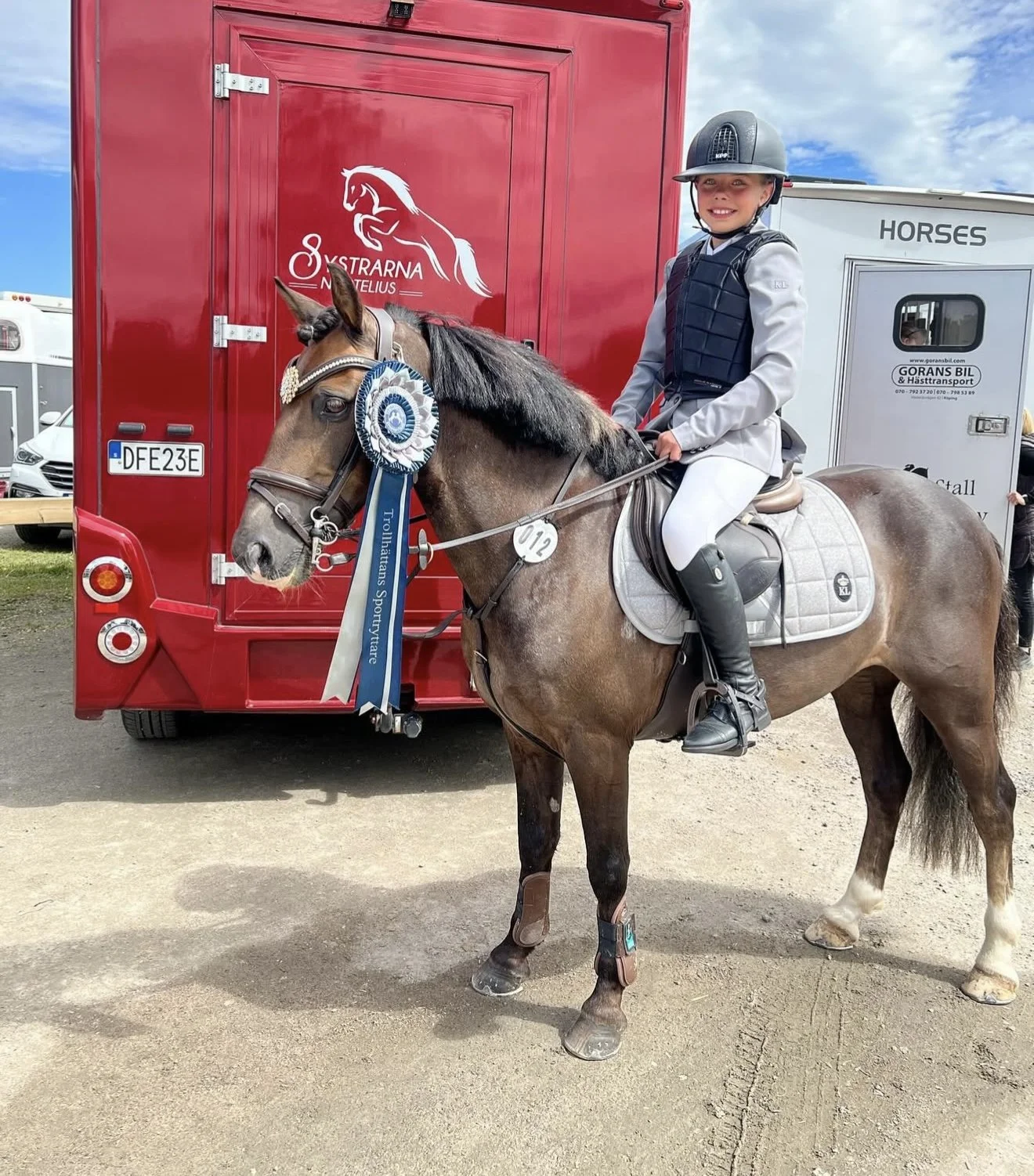 Young girl riding a horse, wearing equestrian attire and a helmet, in front of a red horse trailer with awards attached to the horse's bridle.
