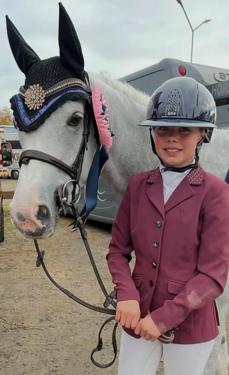 Young girl in riding attire standing next to a gray horse with a decorative bridle and an English riding helmet.