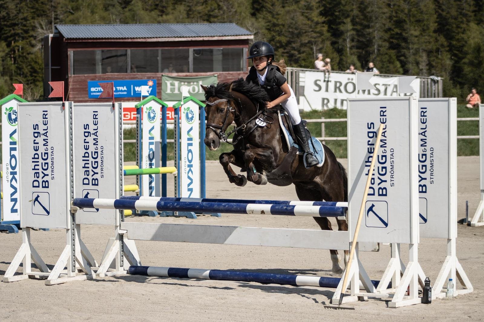 A young female equestrian rider wearing a helmet and black riding attire guides her dark brown horse as they jump over an obstacle during a show jumping event. The course is set on a sandy arena with banners and spectators in the background.