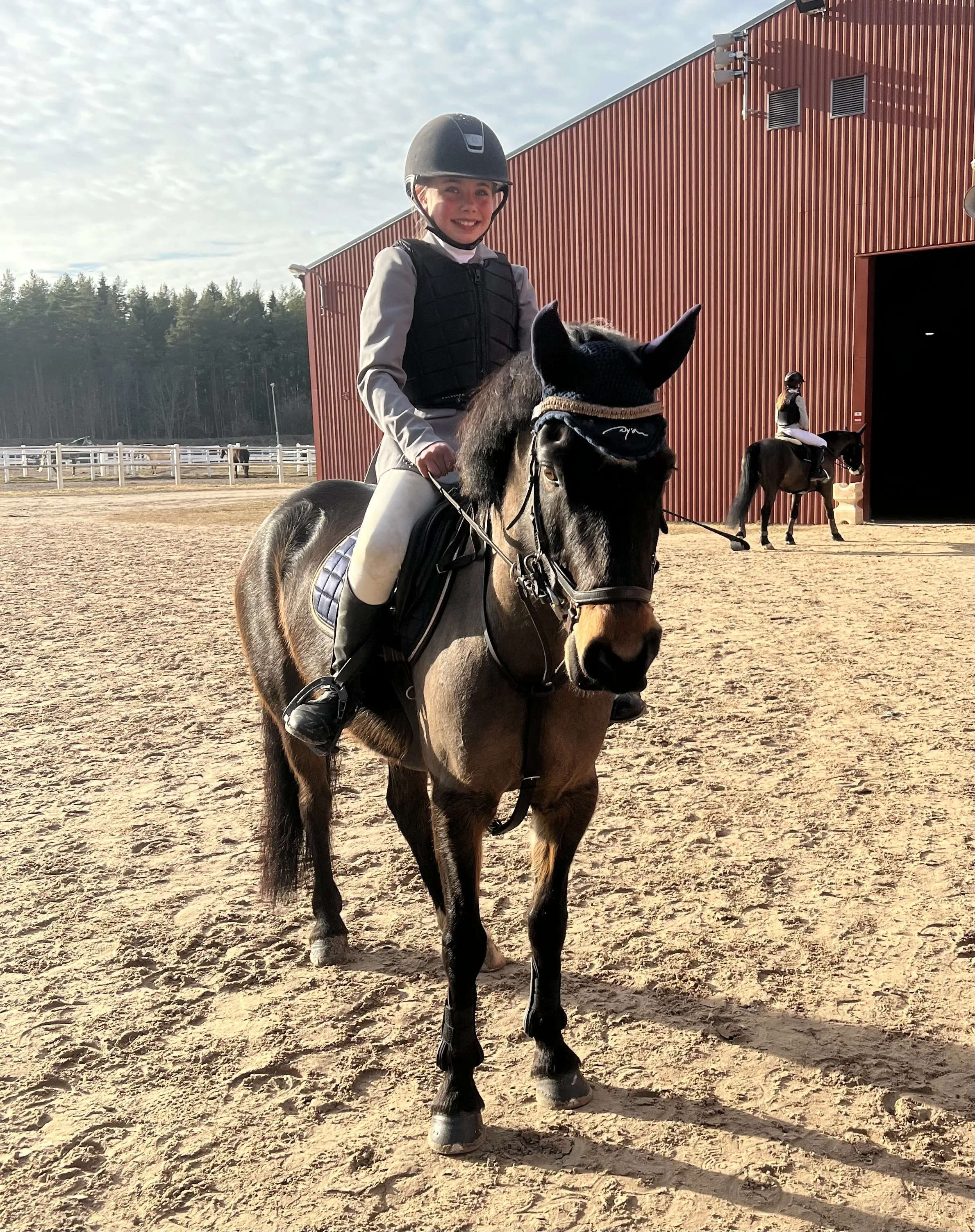 Young girl in riding gear with helmet riding a small brown horse with black mane and tail on a sandy outdoor riding arena. Another rider is seen in the background near a red barn, with a wooded area beyond.