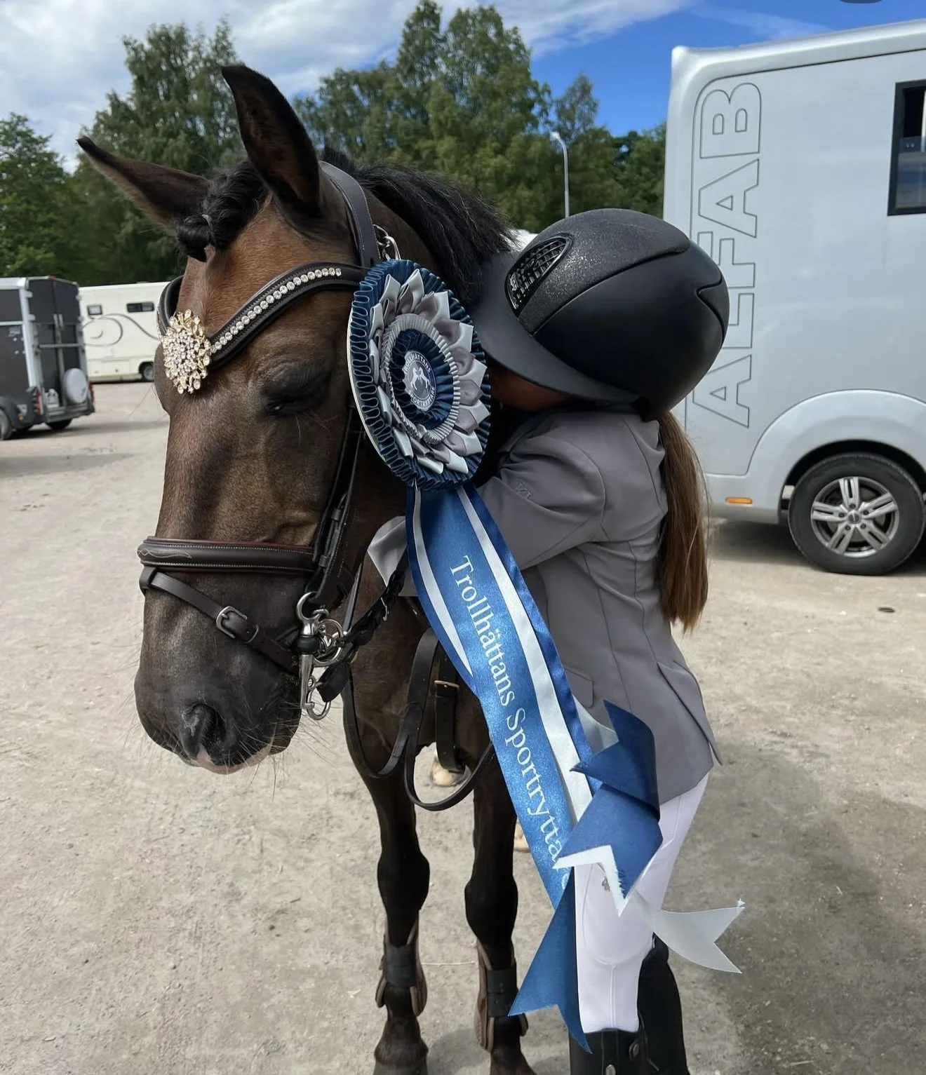 A person wearing a helmet and gray jacket hugging a brown horse decorated with a blue and silver ribbon and rosette, indicating a winning achievement at a riding competition, with trailers and trees in the background.