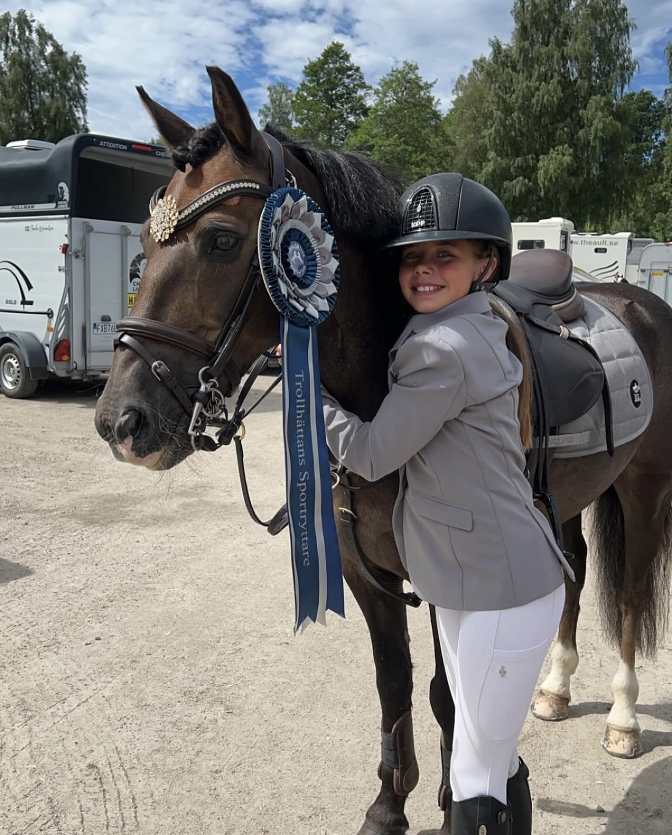 A girl in riding attire and a helmet hugging a horse decorated with a blue and white ribbon and a flower headpiece, standing outdoors on a dirt area with trees and parked trailers in the background.