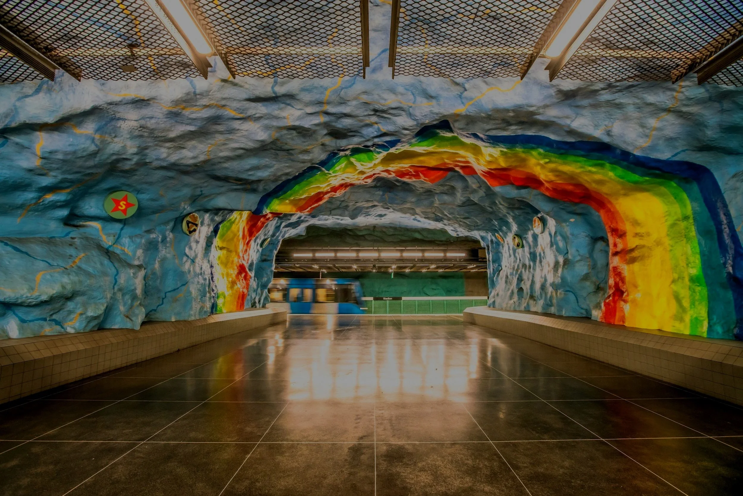 Estación de metro con una cueva decorada con pintura de arcoíris en la pared y techo, y un tren azul pasando por el andén.