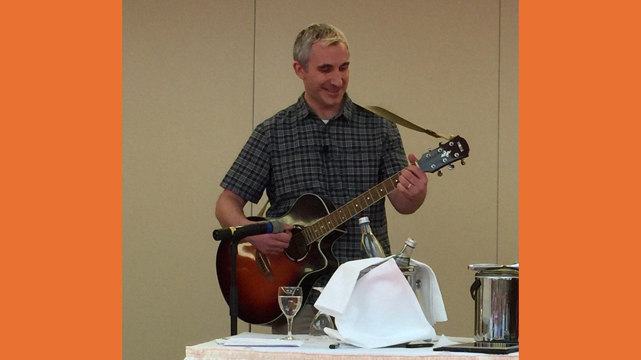 A man in a plaid shirt playing an acoustic guitar at a table with a microphone, a glass of water, and a napkin, smiling.