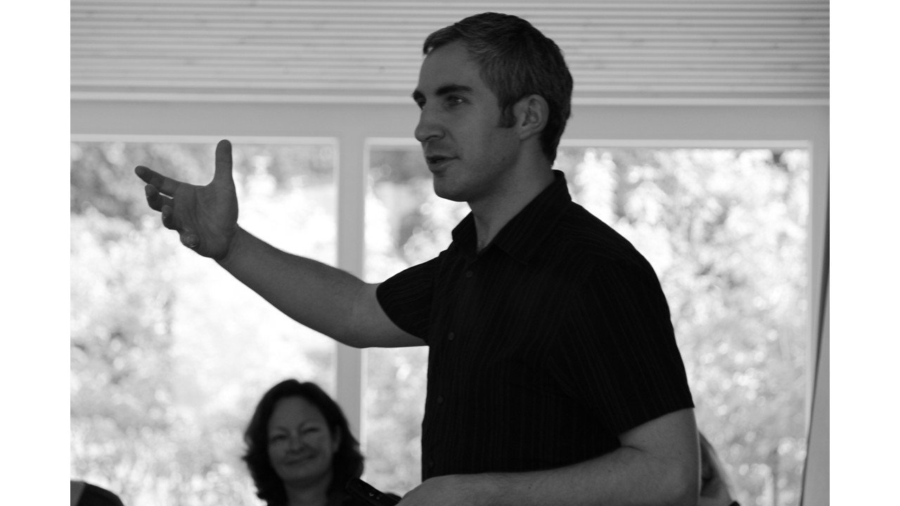 A man gesturing with his right hand while speaking indoors, with a woman smiling in the background.