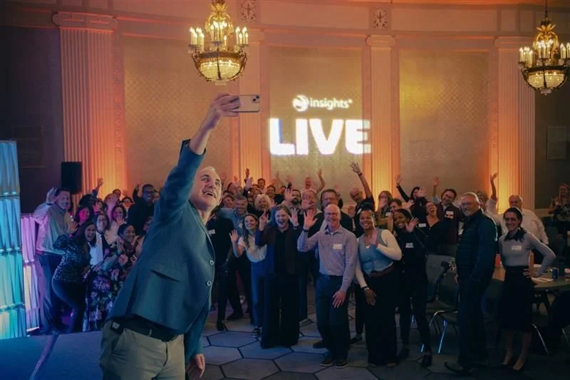 Group of diverse people taking a selfie at a professional event with a large screen displaying 'insights LIVE' in the background, inside a decorated ballroom with chandeliers.