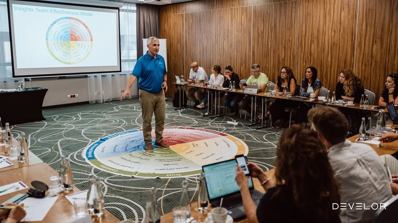 A man in a blue shirt giving a presentation to a group of people sitting around a U-shaped conference table in a room with wooden walls. The presenter stands on a circular diagram on the carpet and points at a projector screen displaying a slide titl
