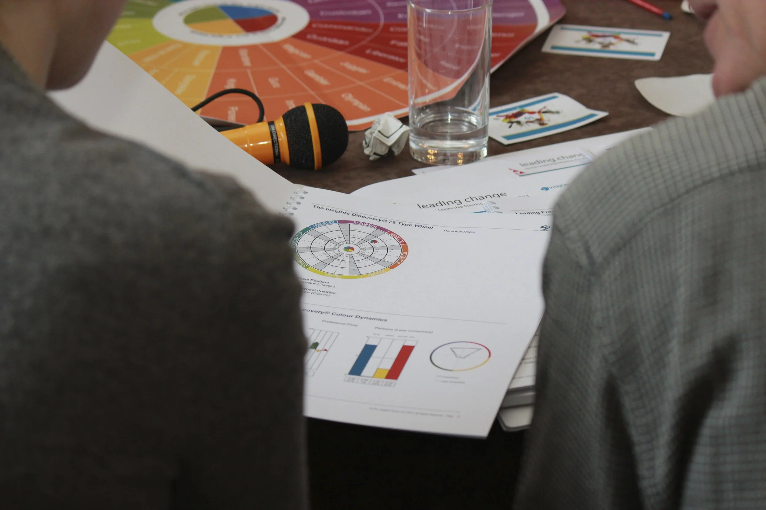 A table with printed charts, a glass of water, a microphone, and business cards, seen from behind two people during a meeting or workshop.