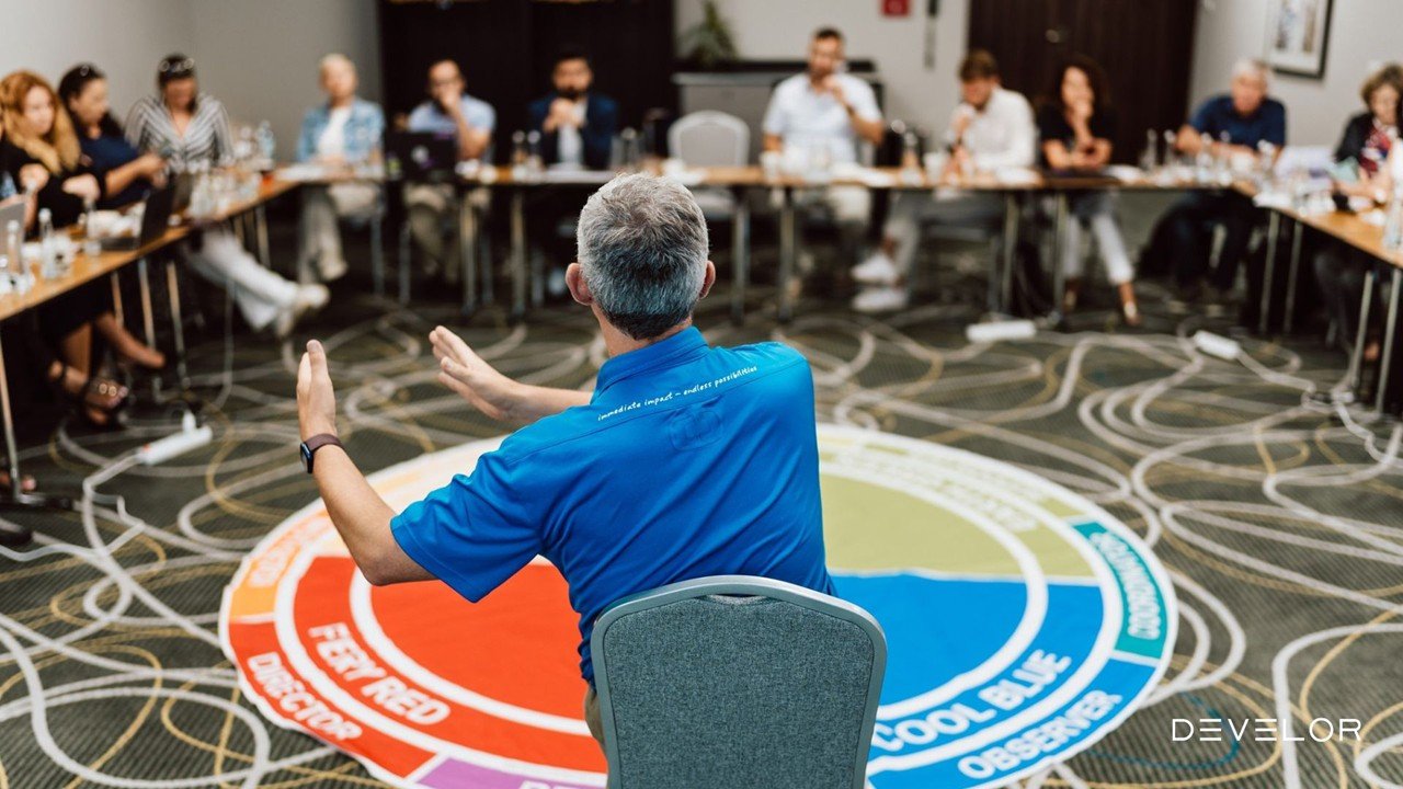 A man in a blue shirt is speaking and gesturing with his hands in front of a seated audience in a conference room. The floor has a colourful circular diagram from Insights Discovery with text, and the audience is listening attentively.