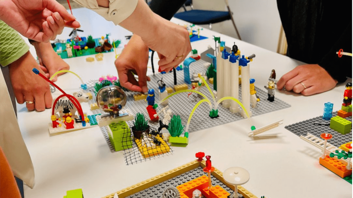 People building a LEGO city on a table with various LEGO structures, figures, and accessories.