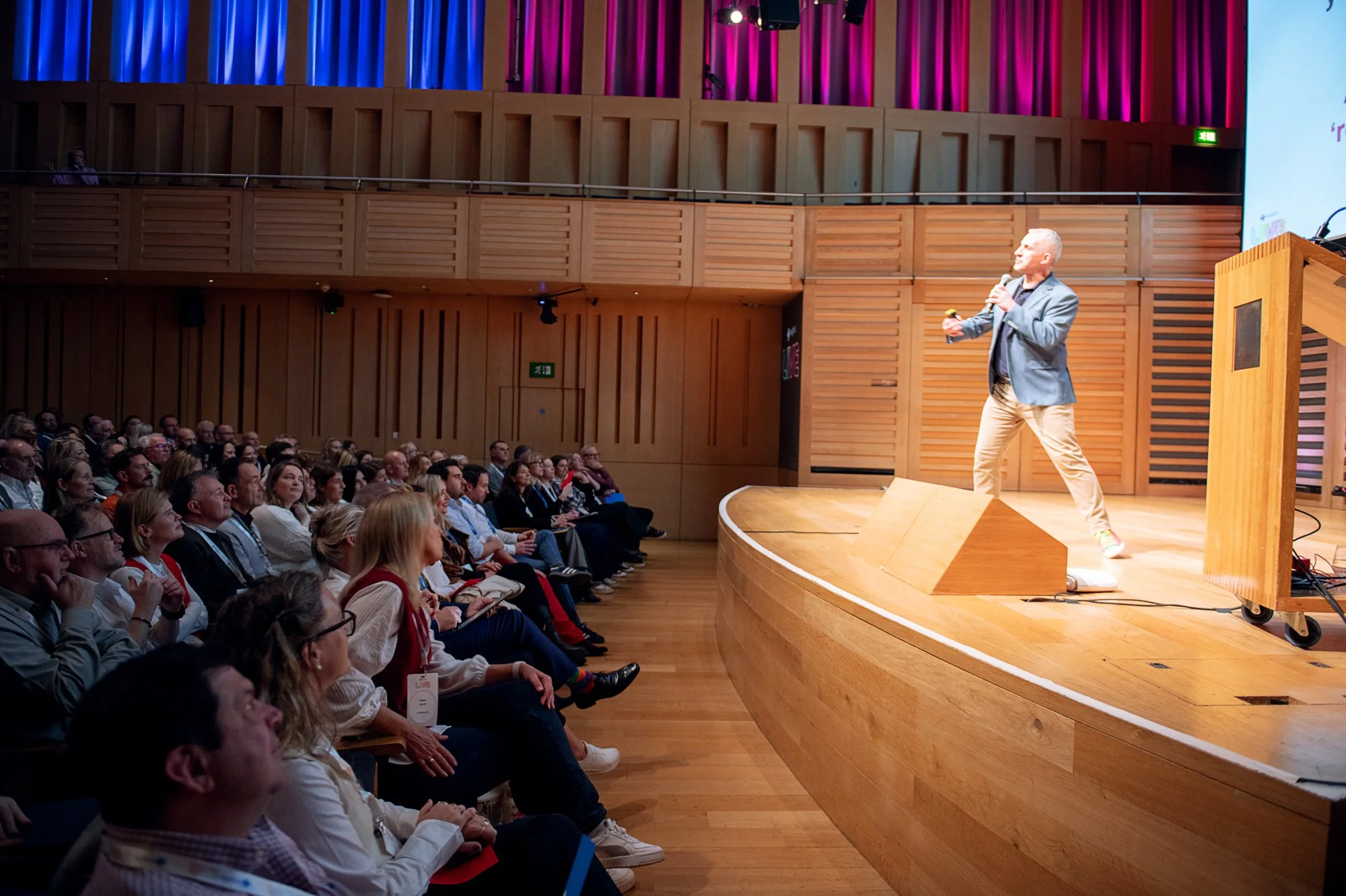 A man giving a presentation on stage at a conference, holding a microphone and gesturing towards the audience, with a large screen behind him and an audience seated in front.