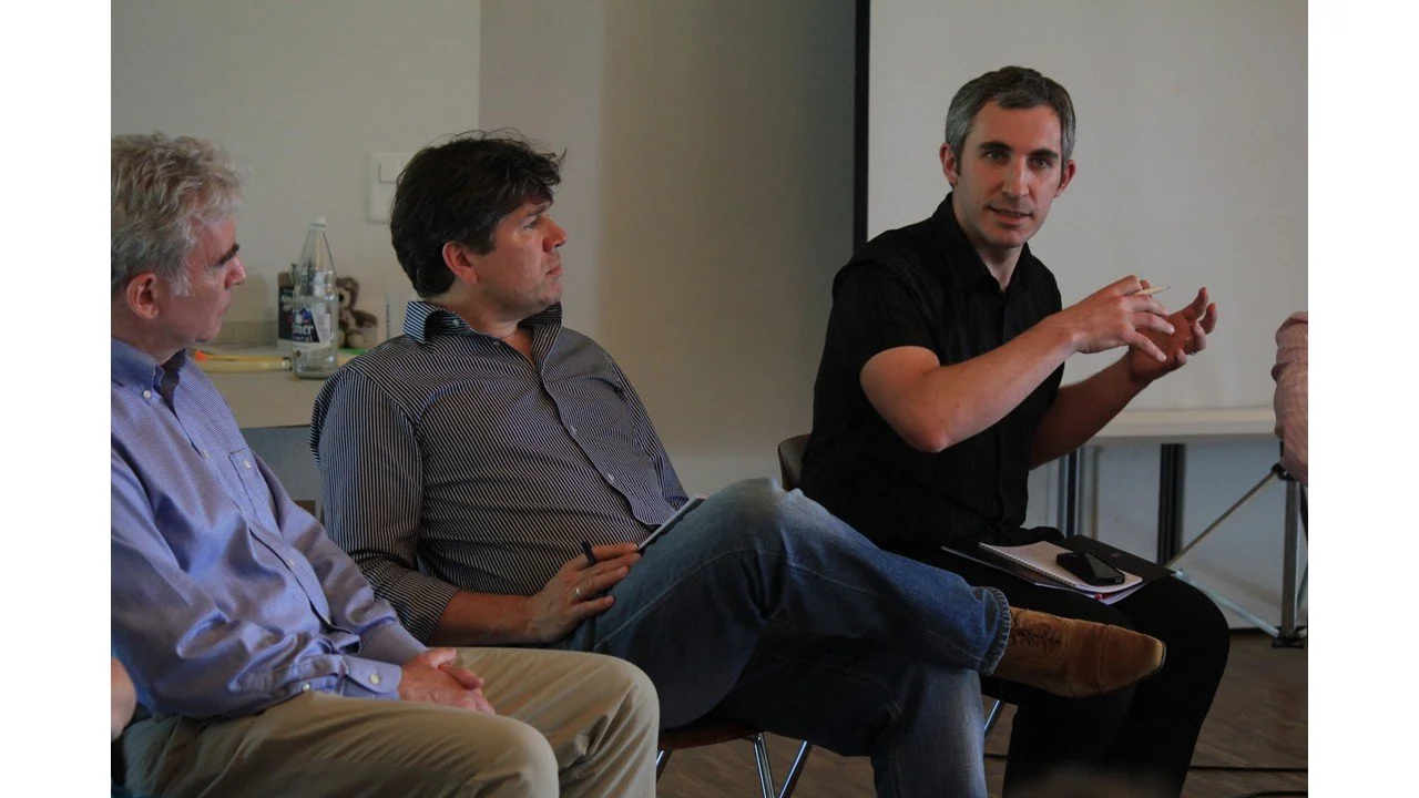 Three men sitting in a meeting or discussion space, one of them actively speaking and gesturing, with a notebook and pen, against a plain background with a table and water bottles.