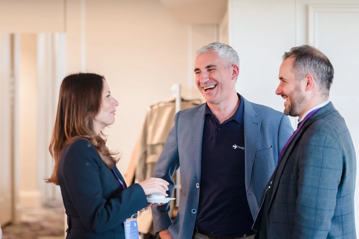 Three professionals are engaged in a friendly conversation, smiling and making eye contact. They are dressed in business attire and appear to be at a conference or networking event.