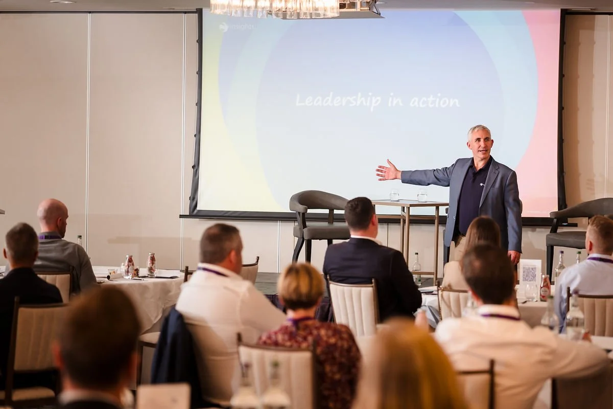 A man in a blazer giving a presentation to a seated audience in a conference room. The presentation slide behind him reads "Leadership in action."
