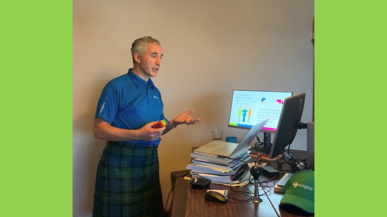 A man in a blue shirt and plaid skirt stands in front of a computer desk, gesturing as he speaks. The desk has a stack of papers, a computer monitor, a laptop, a mouse, and a green cap. There is also a glass of water on the desk.