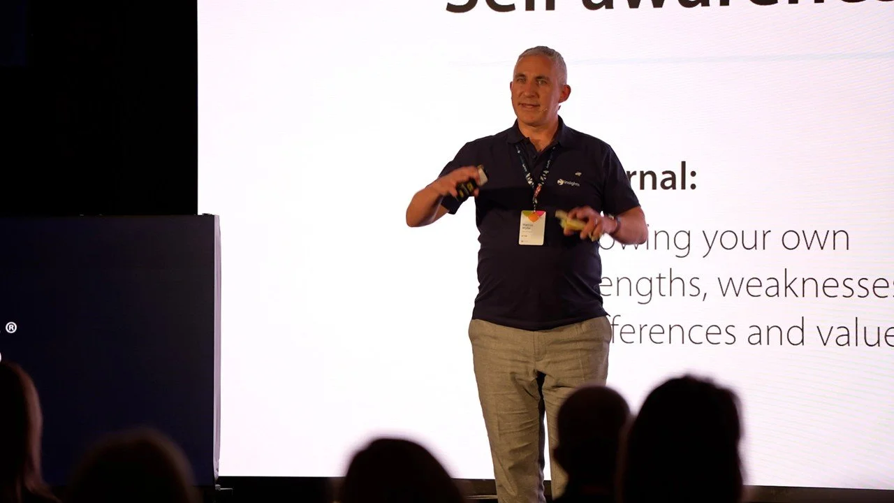 A man giving a presentation on stage, holding a remote and a yellow object, in front of a large screen with text. The audience is visible in the foreground.