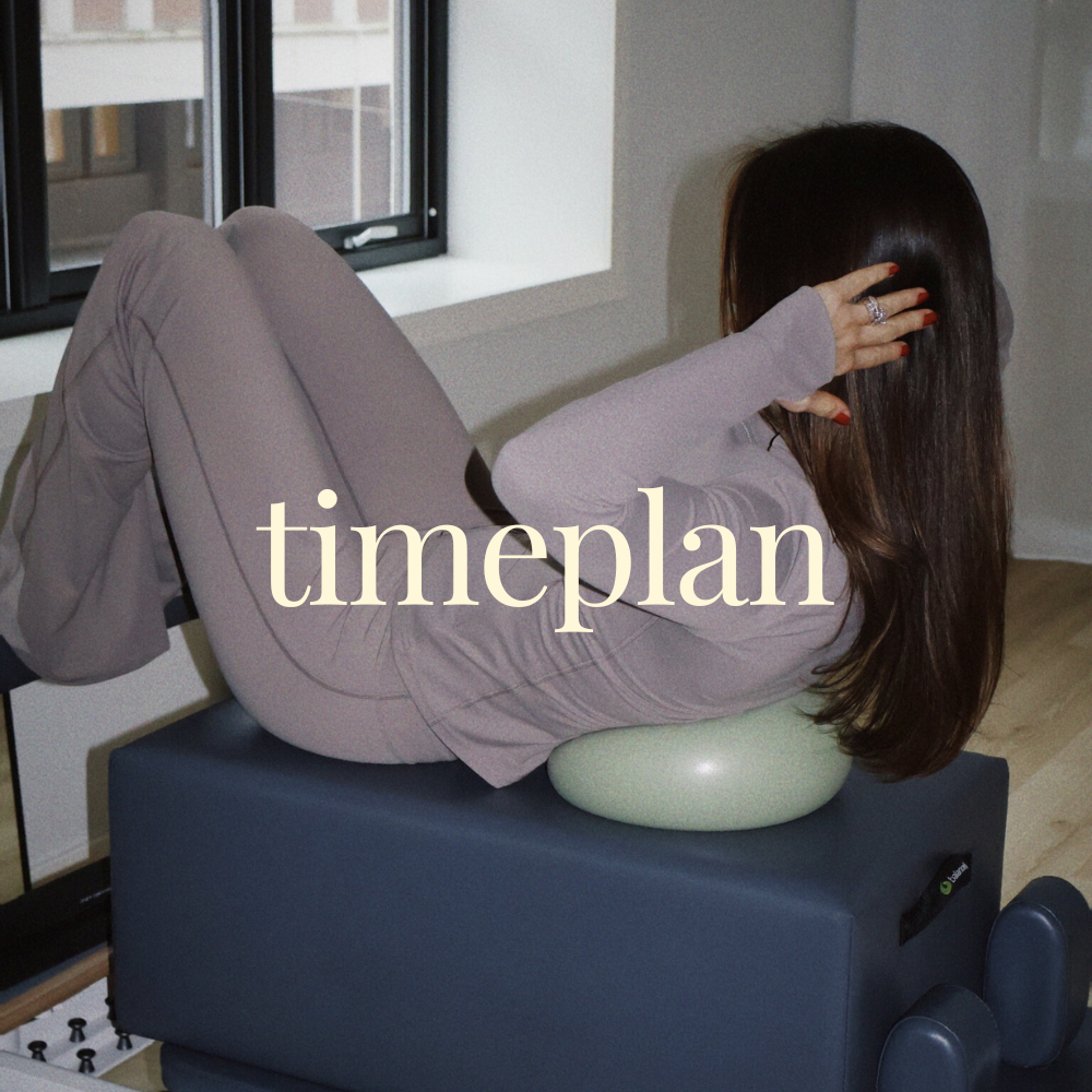 A woman lying on her back on a massage table with her head resting on a foam pillow, near a window with open blinds. The setting appears to be indoors with natural light coming in.