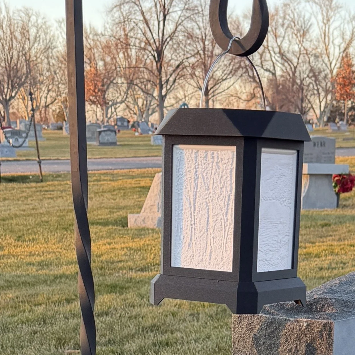 Black lantern with white textured glass panels hanging at a cemetery during sunset.