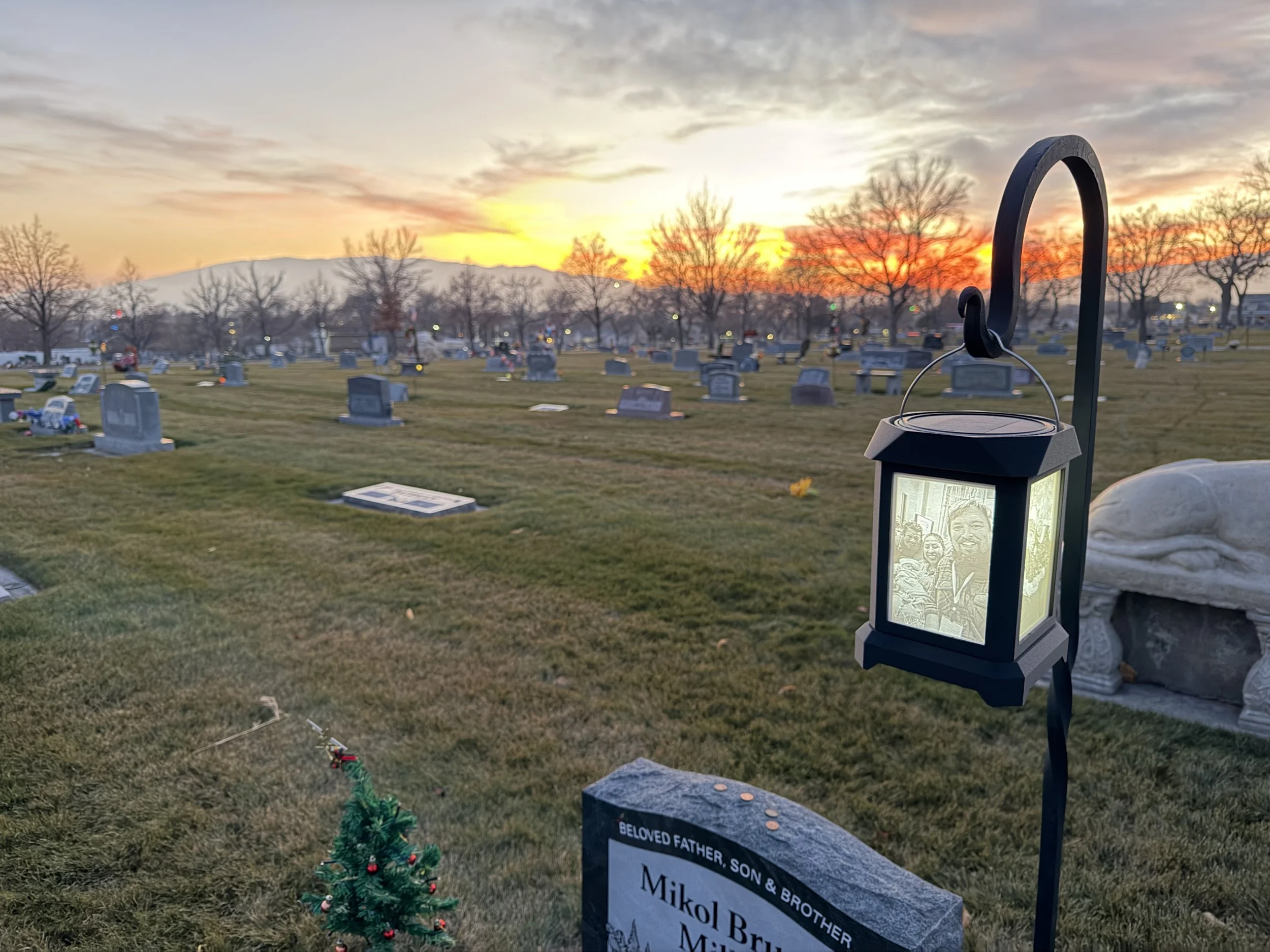 A cemetery at sunset with numerous gravestones and a small, decorated Christmas tree in the foreground. A lantern with a photo of a person inside hangs on a black metal hook near a gravestone inscribed with Miko Bru.