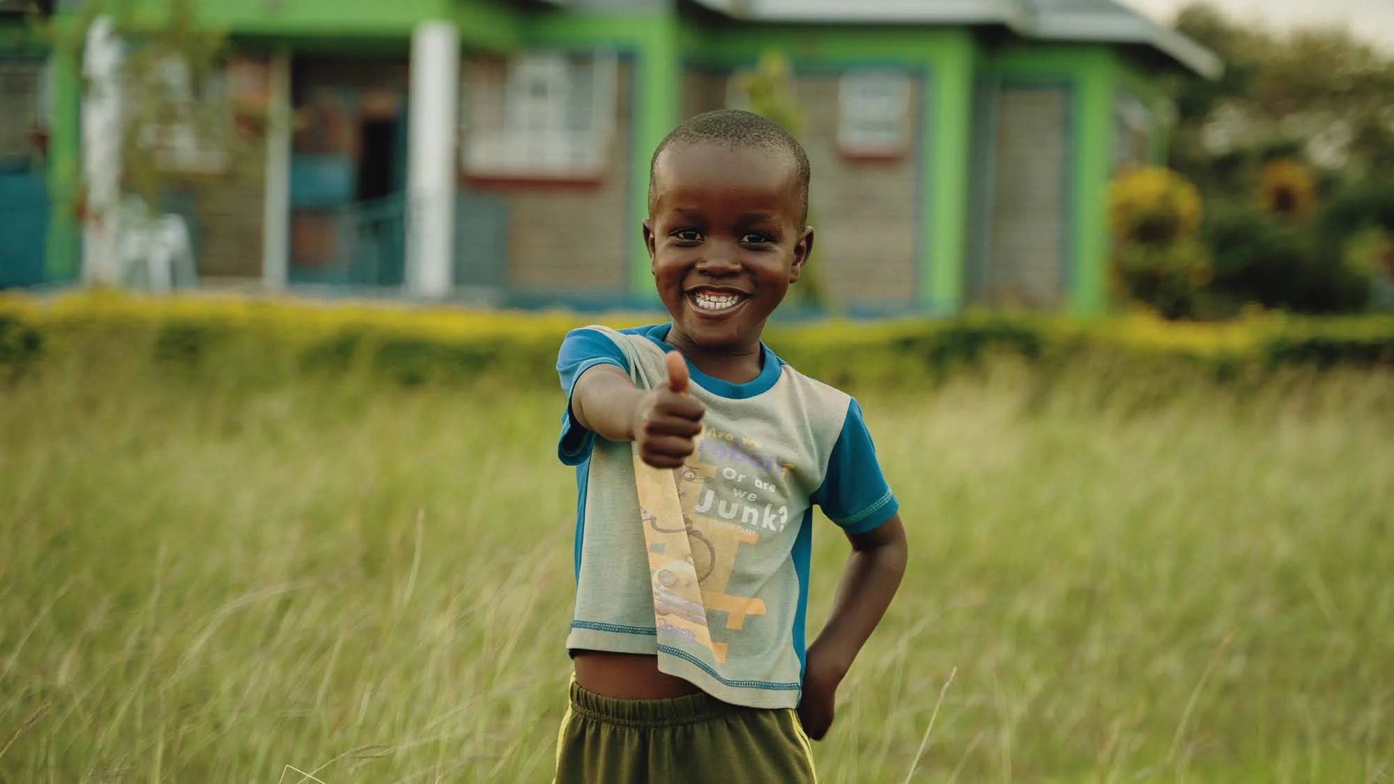 Smiling young boy in a t-shirt and shorts giving a thumbs-up while standing in a grassy field with a colorful house in the background.