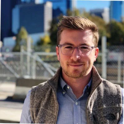 A young man with glasses and short brown hair standing outdoors with city buildings and greenery in the background.