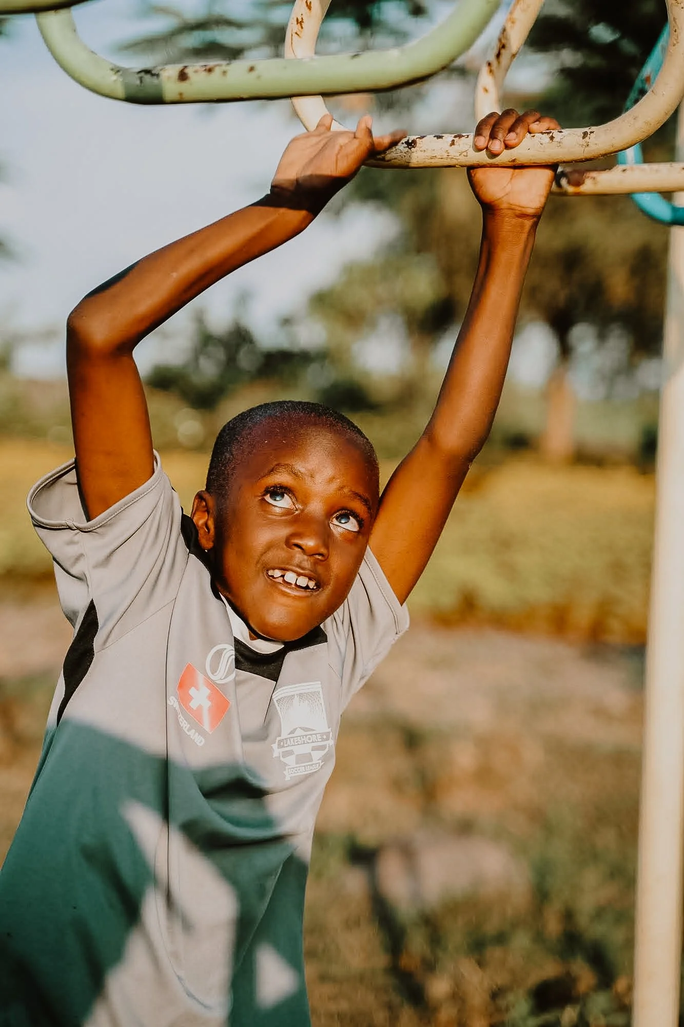 A young boy with short hair hanging from a jungle gym outdoors, with a focused expression on his face.