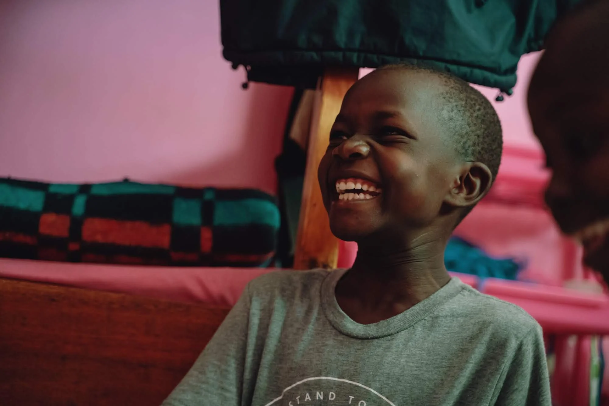 Young boy with a big smile, sitting indoors, with pink walls and a bed with a checkered blanket in the background.