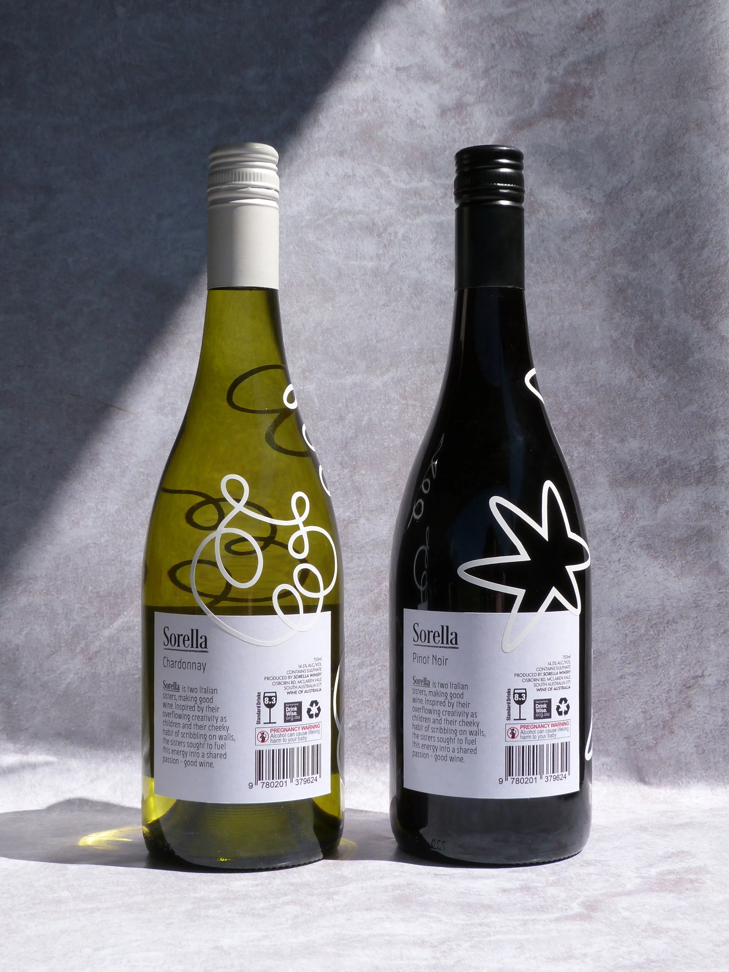 Two wine bottles, one white and one black, placed side by side on a paper surface with a textured gray background. The white wine bottle has a yellow-green tint with a white cap, and the black wine bottle has a black cap. Both bottles have artistic w