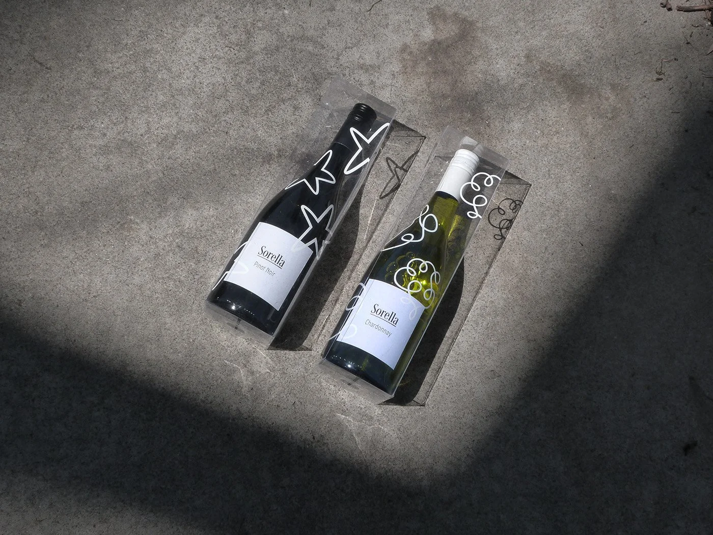 Two wine bottles, one Pinot Noir and one Chardonnay, in transparent gift boxes with black and white abstract designs, placed on a concrete surface with shadows.