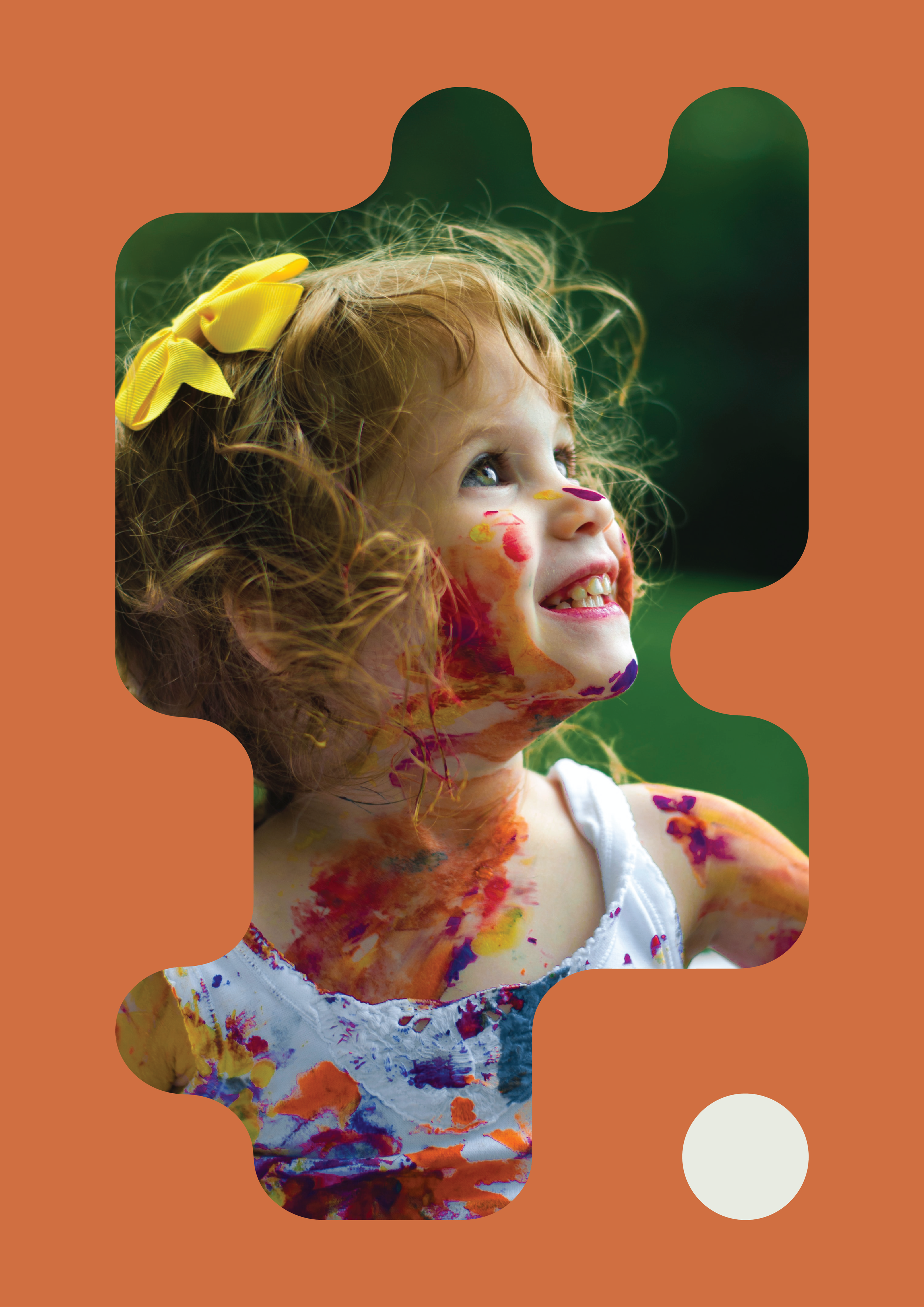 A young girl with curly red hair, smiling and looking upward, has colorful paint on her face and torso, and wears a white dress with paint smudges, outdoors with a green blurred background.