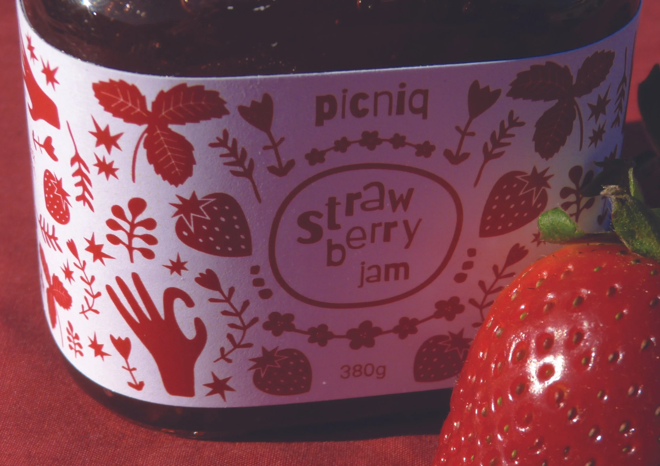 A jar of strawberry jam labeled 'Strawberry Jam' with decorative strawberries, leaves, and hearts on the label. A fresh strawberry is placed next to the jar.