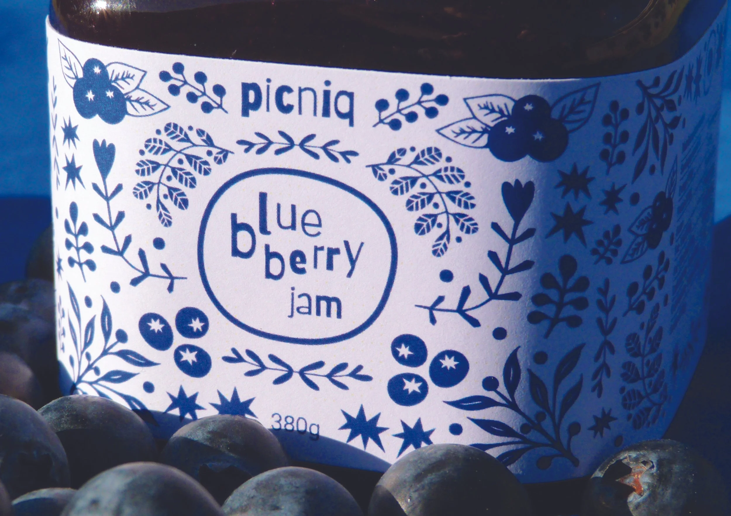 A jar of blueberry jam with a festive, blue and white label featuring blueberry illustrations and decorative foliage, surrounded by fresh blueberries.