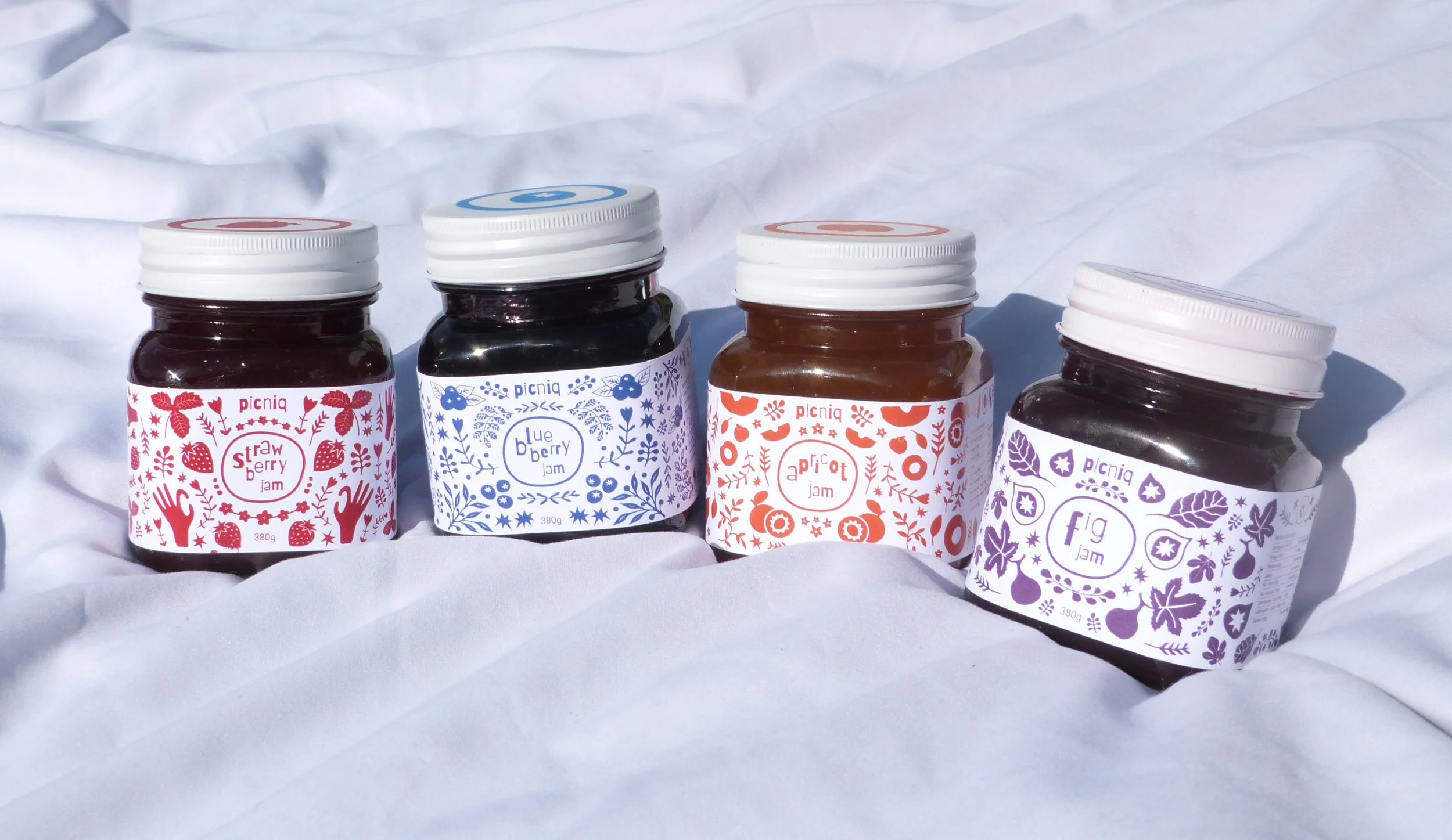 Four jars of fruit jam with colorful labels for strawberry, blueberry, apricot, and fig, placed on a white fabric surface.