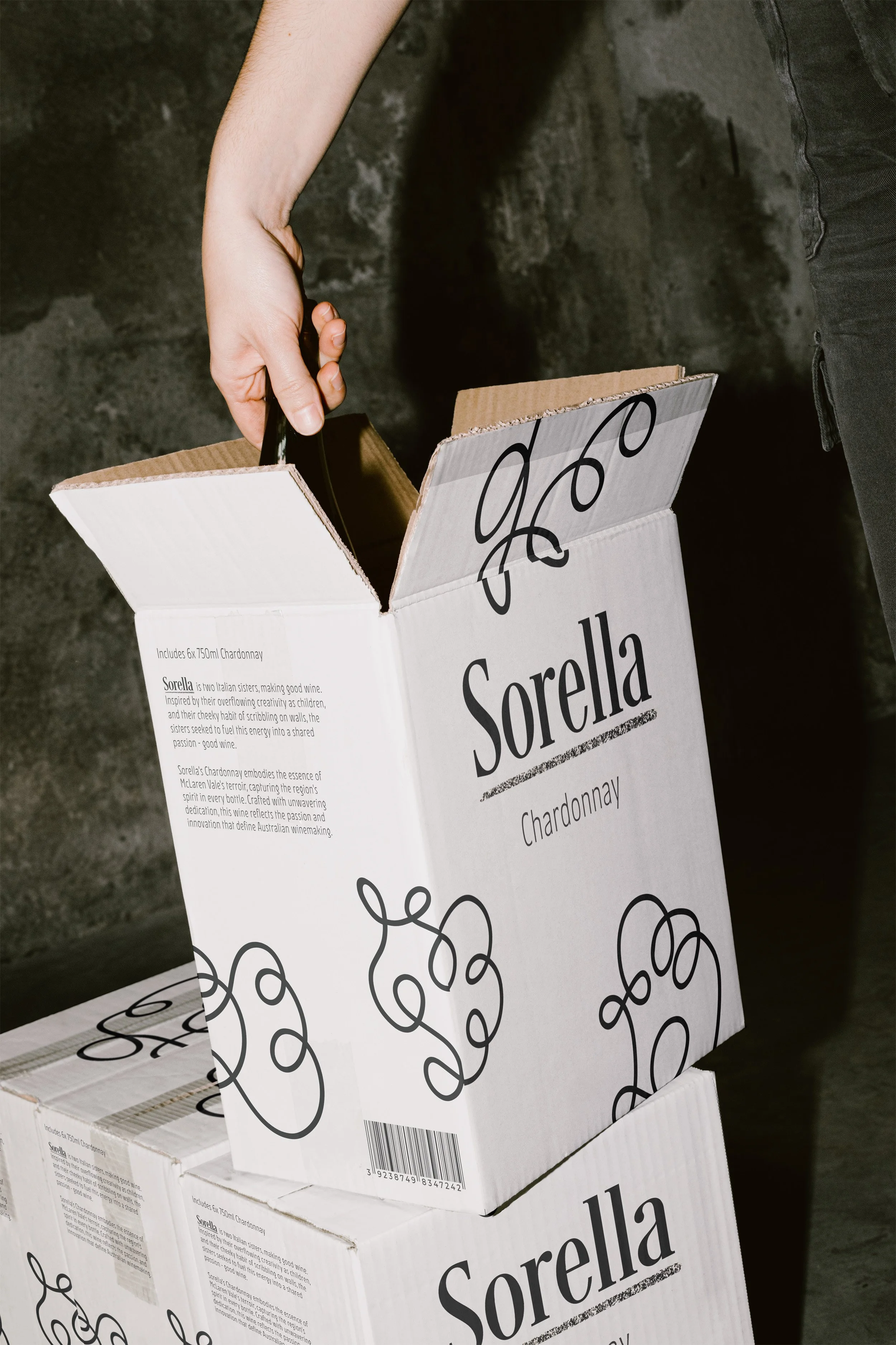 Person opening a box of Sorella Chardonnay wine bottles, with additional boxes stacked below.