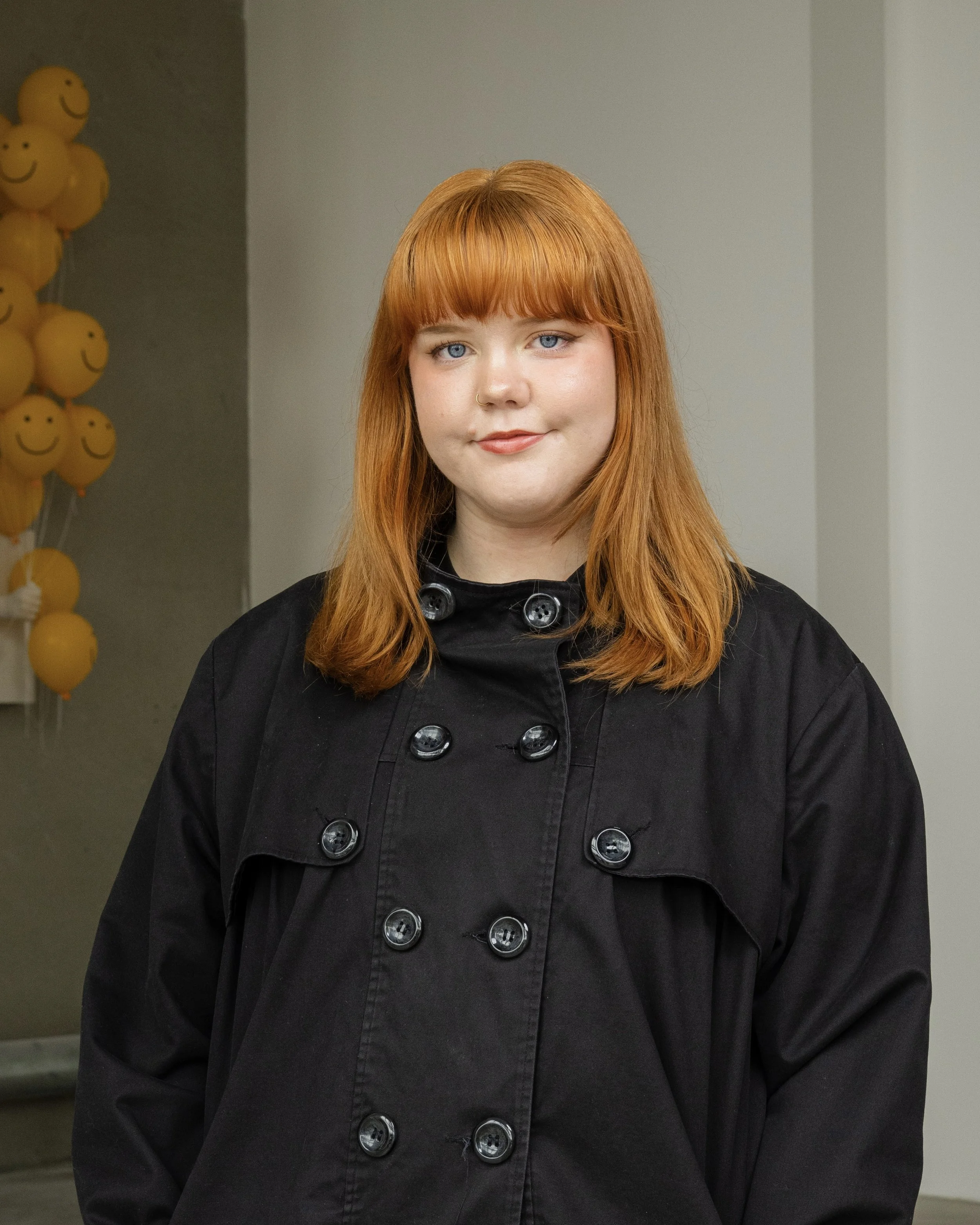 A young woman with red hair, blue eyes, and a nose ring, wearing a black coat with large buttons, standing indoors with yellow smiley face balloons in the background.