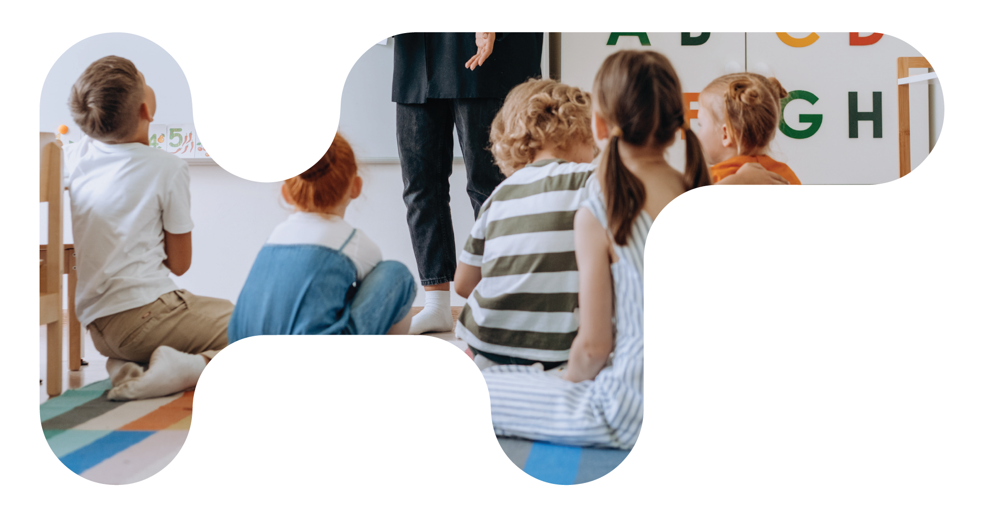 A group of young children sitting cross-legged on the floor of a classroom, listening to a teacher who is standing nearby. The classroom has colorful alphabet letters on the wall and a whiteboard.