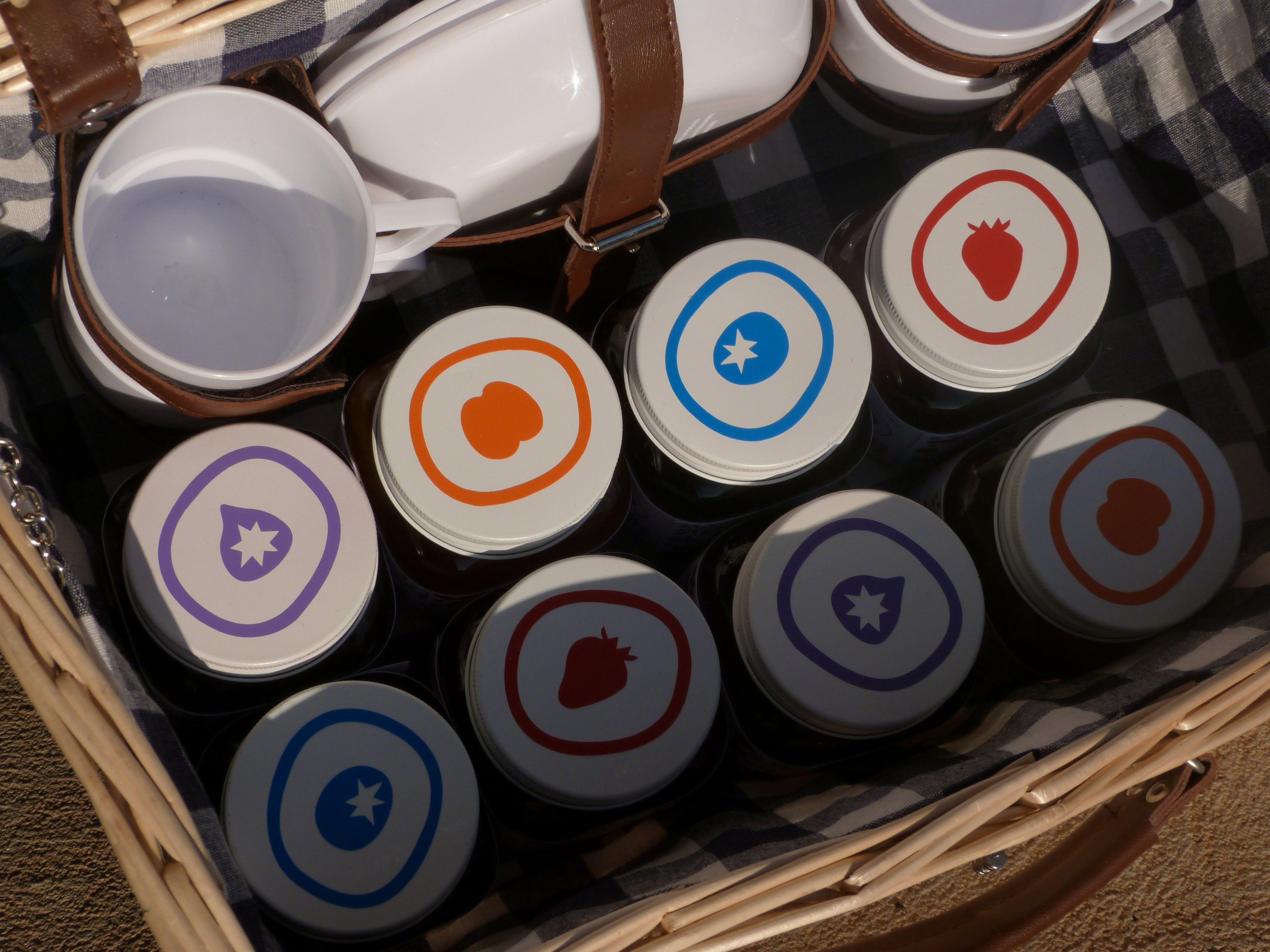 A basket filled with small jars of jam or honey, each with a colorful fruit-themed label on the lid, placed on a checkered cloth.