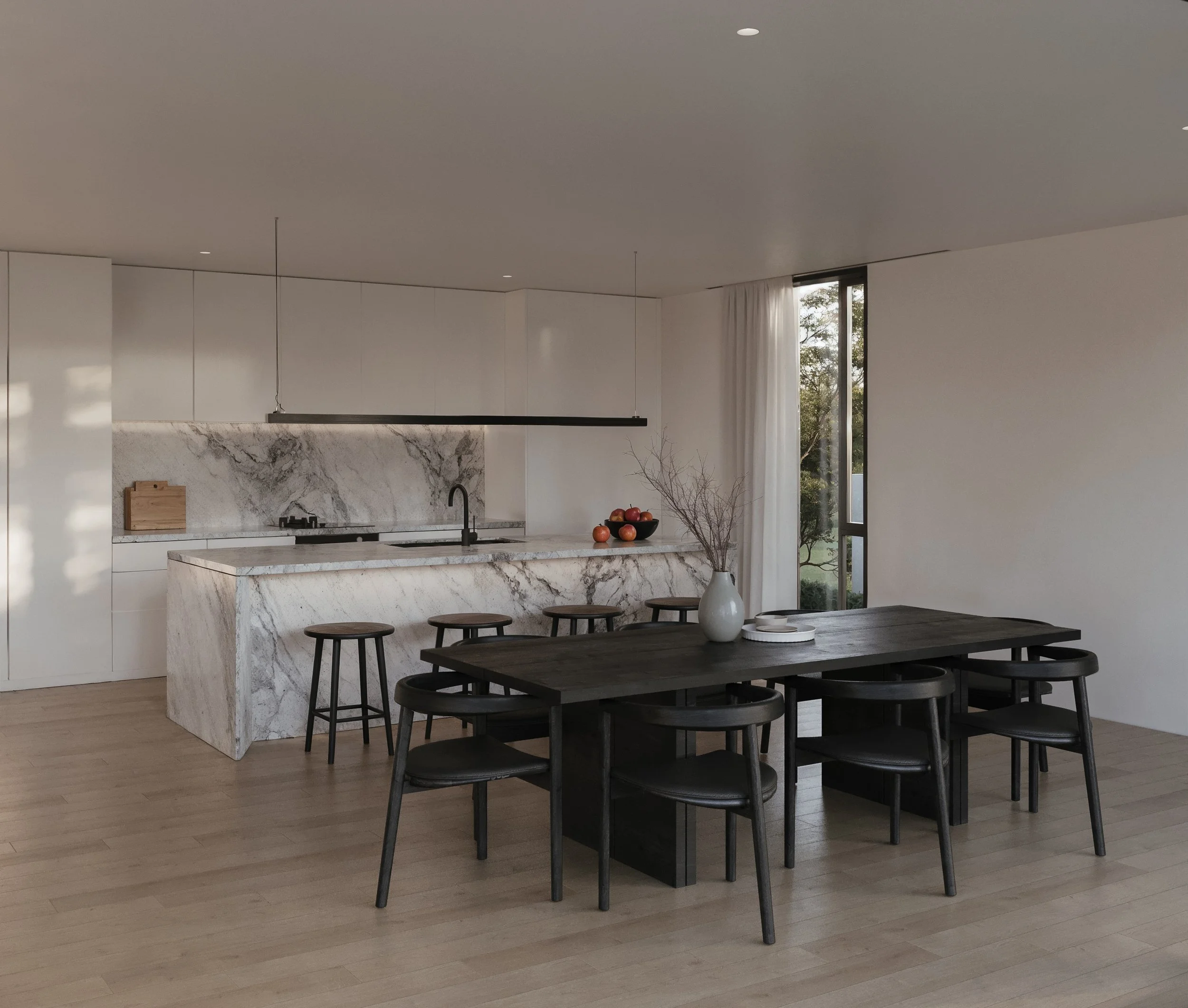 Modern kitchen with white cabinetry, marble island, black stools, wooden dining table with black chairs, vase with branches, and large window with white curtains.