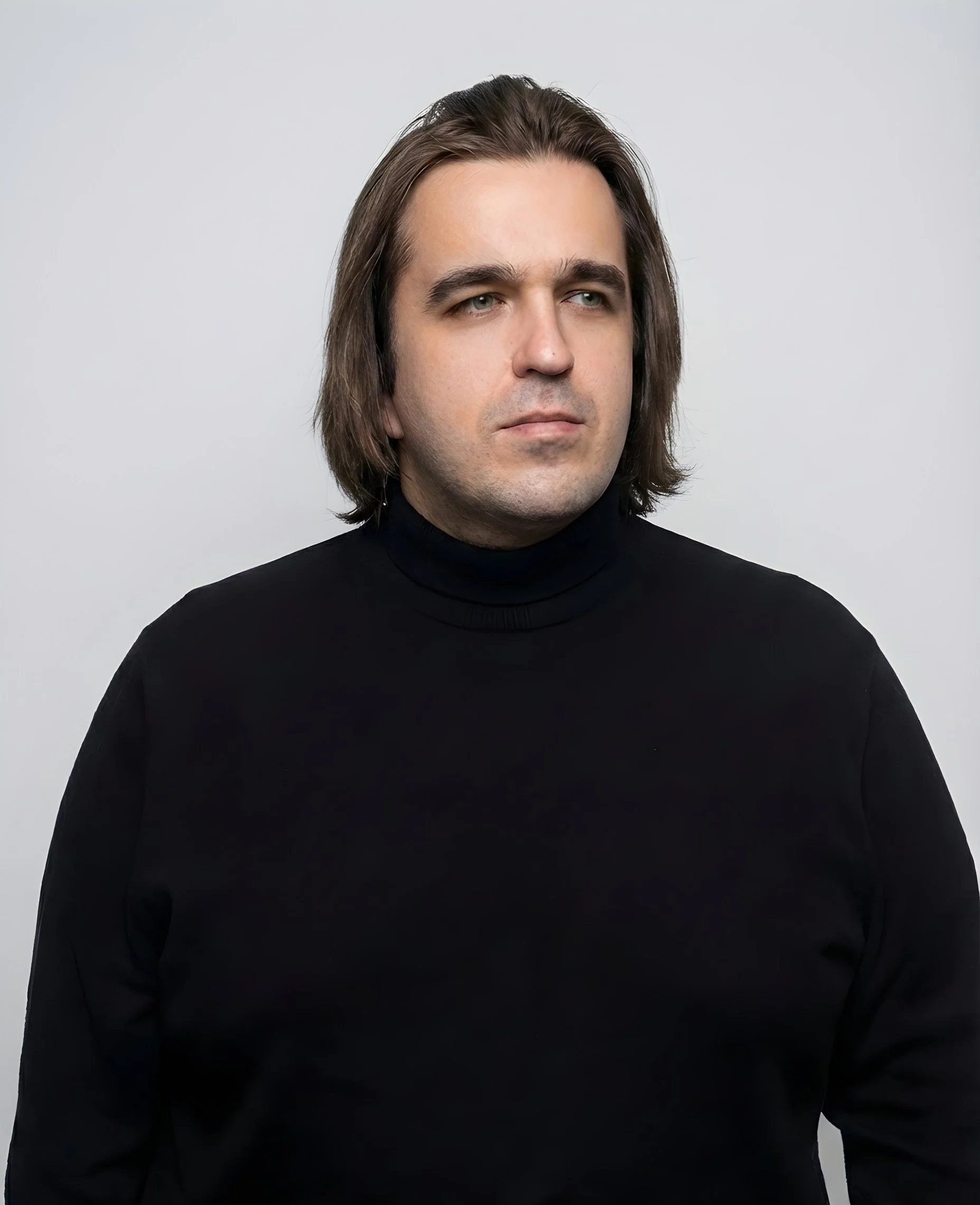 A man with shoulder-length brown hair, wearing a black turtleneck, looking to his right against a plain light gray background.