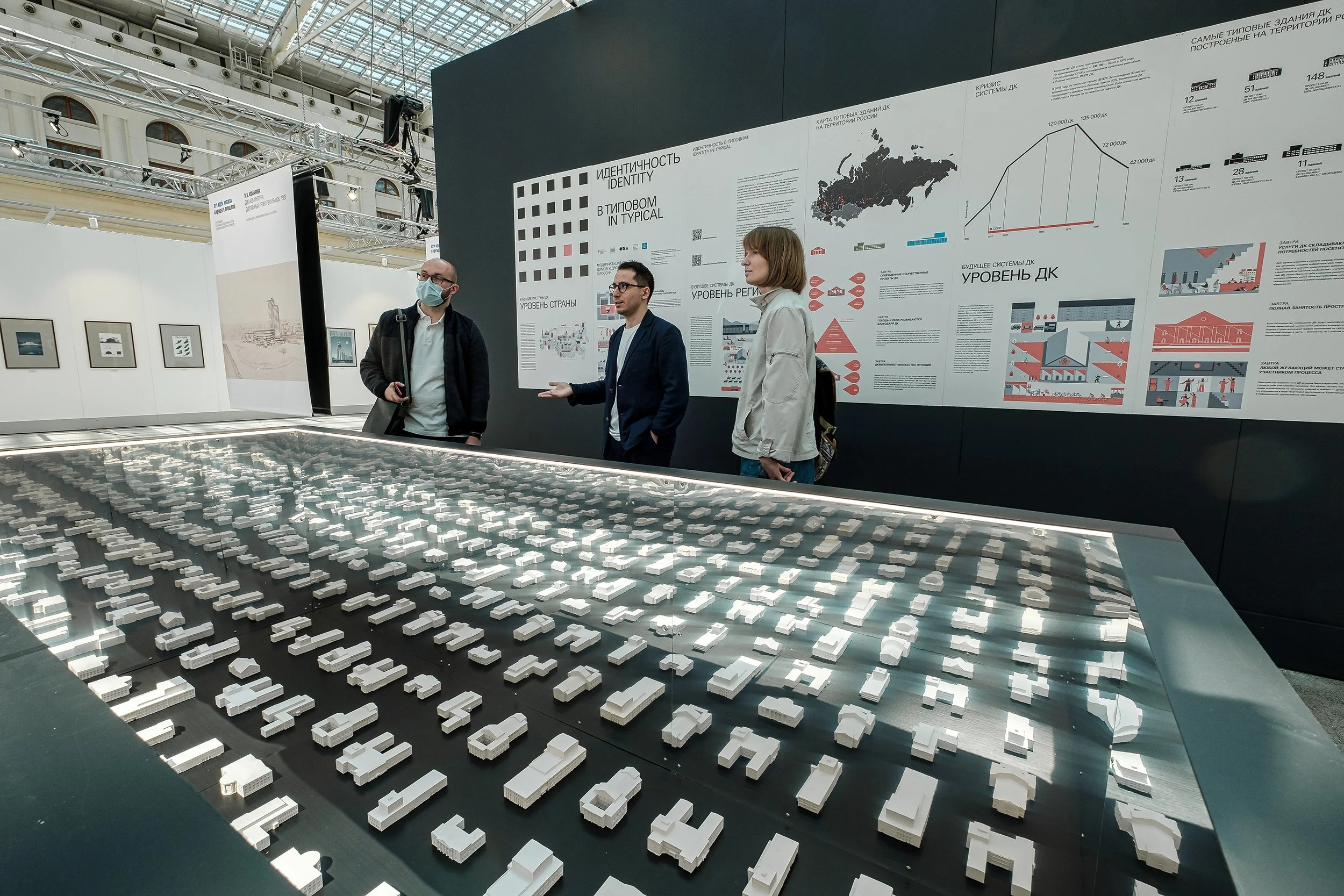 Museum exhibit with a detailed architectural model of a city, behind a black wall with informational posters about Russia's identity, typical buildings, and future infrastructure plans, three people engaging in conversation.