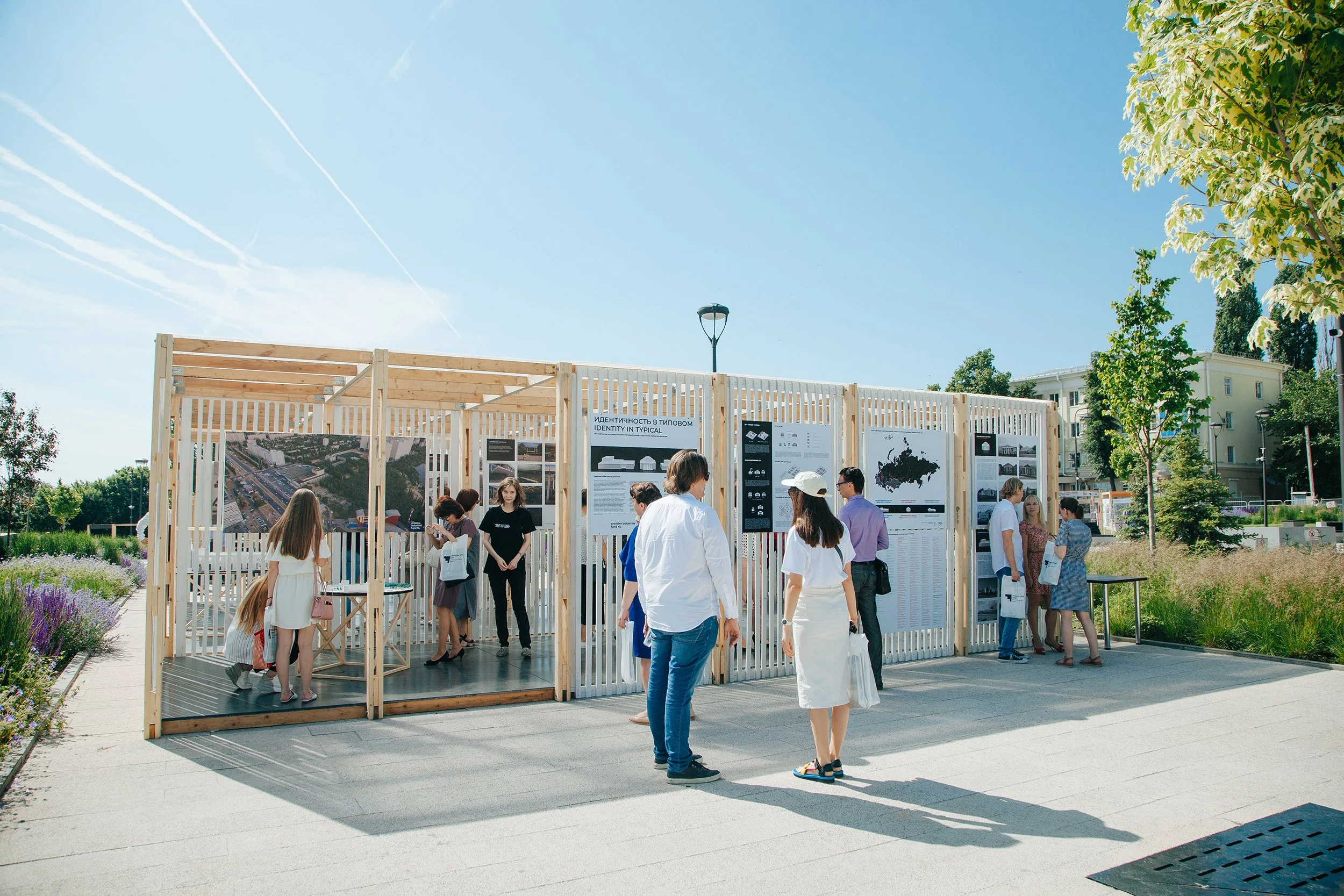 Outdoor exhibition with people viewing posters and displays on wooden panels under a clear blue sky.